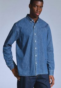 Camicia in denim blu scuro con bottoni, dotata di colletto, maniche lunghe e bottoni bianchi. Il tessuto è morbido e con una texture liscia.