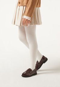 Dunkelburgunderfarbene Lackleder-Loafer mit flacher Sohle, getragen mit weißen Strumpfhosen und einem gefalteten beigen Rock, kombiniert mit einem strukturierten Strickpullover.