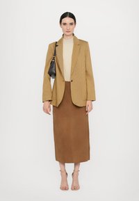 Kahbrown pinstriped blazer yli beige toppi, yhdistettynä ruskeaan midi-hameeseen. Malli pitää pientä mustaa käsilaukkua ja käyttää beigejä nilkkasandaleita.