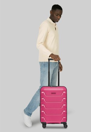 Mann in beige-langärmlichem Hemd und Jeans steht und hält den ausgezogenen Griff eines pinkfarbenen Hartschalenkoffers mit vier Rollen.