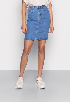 Vrouw die een blauwe denim minirok, een beige ingesopt overhemd en witte chunky sneakers draagt, staand tegen een effen achtergrond.
