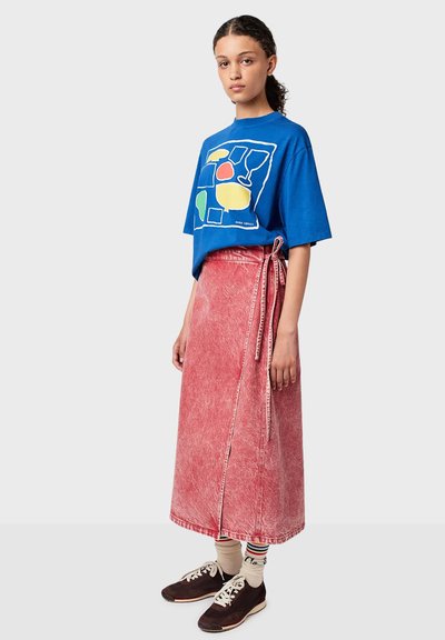 Jeune femme portant un t-shirt graphique bleu, une jupe portefeuille rouge délavée, des chaussettes rayées et des baskets marron, se tenant devant un fond uni.