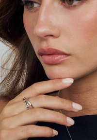 Närbild av en modells ansikte med mjuka hudtoner, fylliga nakna läppar, naturliga naglar och en silverring på fingrarna som vilar på hakan.