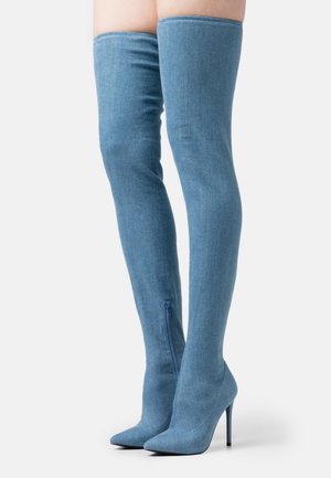 Stivali blu in denim alti fino alla coscia con punte a punta, tacchi a stiletto e cerniere laterali indossati su gambe slanciate su uno sfondo bianco.
