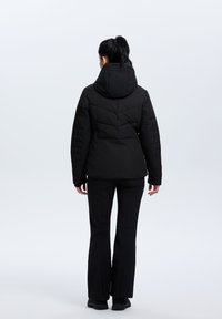 Chaqueta negra acolchada con capucha, con un diseño acolchado y puños elásticos, llevada con pantalones y zapatos negros, vista desde atrás.