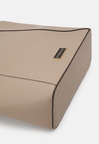 Beige kožená taška obdélníkového tvaru, s černým lemováním a zlatou logovou destičkou na jedné straně. Hladký povrch, minimalistický design.