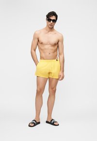 Άνδρας που φοράει μαύρα γυαλιά ηλίου, κίτρινα μαγιό με κορδόνι και μαύρα σανδάλια Moschino, στέκεται μπροστά σε ένα απλό ανοιχτό φόντο.