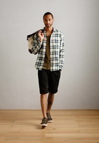 Koszula w kratkę, zapinana na guziki, w zielono-białe wzory, brązowy dzianinowy podkład, czarne szorty, szare sneakersy. Styl casualowy, z torbą na ramię.