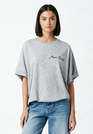 Jonge vrouw met steil zwart haar, die een loszittend grijs T-shirt met de tekst "Mira Paris" en een blauwe spijkerbroek draagt, staand tegen een effen achtergrond.