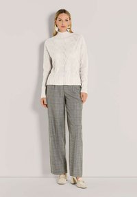 Frau mit langen blonden Haaren, die einen weißen gemusterten Strickpullover, graue karierte Hose im weit geschnittenen Stil, cremefarbene Blockabsatzslipper und geometrische Ohrringe trägt.