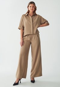 Tan coordinated outfit featuring a short-sleeve button-up shirt and wide-leg trousers. Smooth fabric with a relaxed fit and pleated detailing.