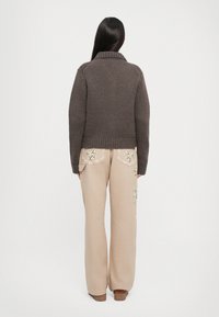 Maglione marrone a maglia con colletto a coste, abbinato a pantaloni beige larghi con tasche ricamate a fiori. Entrambi presentano una finitura testurizzata.