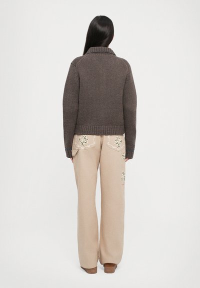 Brauner gestrickter Pullover mit einem gerippten Kragen, kombiniert mit weiten beigen Hosen, die über bestickte Taschen mit Blumenmotiven verfügen. Beide Teile haben eine strukturierte Oberfläche.