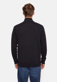 Svart stickad tröja med hög krage, långa ärmar och ribbade muddars, bärs med blå jeans. Slät textur, åtsittande design, inga synliga mönster.