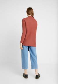 Maglione a coste rosso mattone con maniche lunghe, spacchi laterali e collo alto, abbinato a jeans cropped azzurri e scarpe nere.