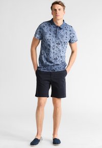 Navy blue polo shirt with floral print, three-button placket, paired with dark shorts and casual slip-on shoes with a striped pattern.