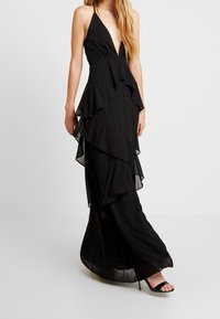 Czarna sukienka maxi z głębokim dekoltem w kształcie litery V, warstwowe falbany spływające z przodu oraz przezroczysty dół sukienki. Materiał wydaje się lekki i zwiewny.