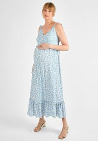 Rochie de maternitate florală de culoare albastru deschis, cu tiv ondulat, bretele ajustabile și material moale. Modelul își mângâie delicat burtica.