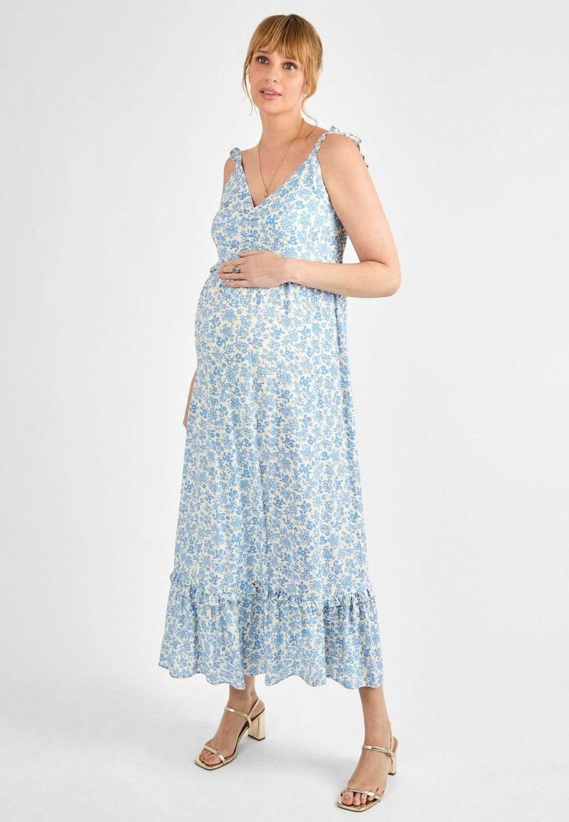 Rochie de maternitate florală de culoare albastru deschis, cu tiv ondulat, bretele ajustabile și material moale. Modelul își mângâie delicat burtica.