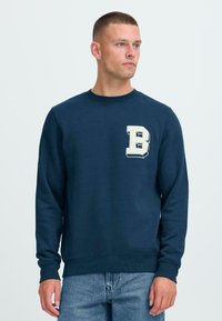 Mann iført en marineblå genser med hvit bokstav "B" på brystet og lyseblåjeans, står mot en ensfarget lys bakgrunn.