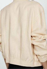 Widok z tyłu osoby noszącej luźną, jasnobeżową kurtkę z gumkami na mankietach i w pasie, w zestawieniu z czarnymi spodniami.