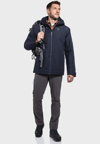 Marine waterdichte jas met capuchon, grijze broek en zwarte wandelschoenen. Inclusief rugzak en heeft een rits en logo-details.