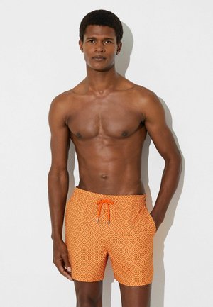 Skiløsbåret mand stående mod en hvid væg iført orange badeshorts med mønster og snøre, den ene hånd i lommen.