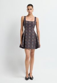 Jeune femme portant une robe sans manches à motifs, évasée et ajustée, et des talons aiguilles noirs, se tenant devant un fond blanc uni.