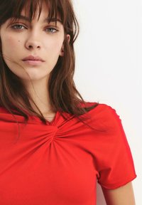 Rode top van lichtgewicht stof, met een knoopdetail bij de halslijn. Korte mouwen en een gladde textuur met zachte plooien.