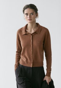 Bruin vest van een wolmengsel met een kraag, knopen aan de voorkant en geribde manchetten. Gecombineerd met zwarte broek staat het model tegen een lichte achtergrond.