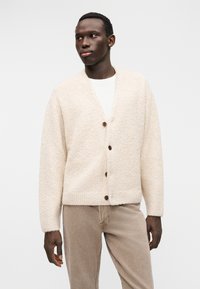 Πλεκτή ζακέτα εκρού χρώματος από υφασμάτινη υφή, με λαιμόκοψη σε σχήμα V, πέντε ξύλινα κουμπιά και μακριά μανίκια, συνδυασμένη με ανοιχτό καφέ παντελόνια.