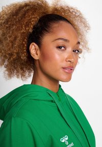 Zielona bluza z zamkiem błyskawicznym i kieszenią kangurzą, z logotypem po lewej stronie. Gładki materiał, regulowany kaptur ze sznurkiem oraz ściągacze przy mankietach.