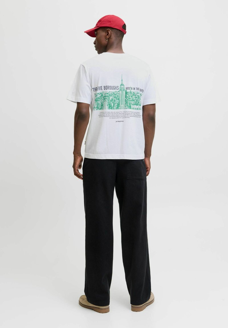 White t-shirt featuring green NYC skyline graphic on the back, paired with black wide-leg pants and a red cap. Simple, casual design.