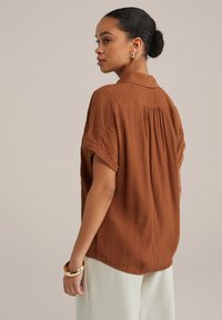 Camicia marrone a maniche corte con un motivo testurizzato, colletto a bottoni e dettagli plissettati sul retro. Indossata con pantaloni bianchi e bracciale d'oro.