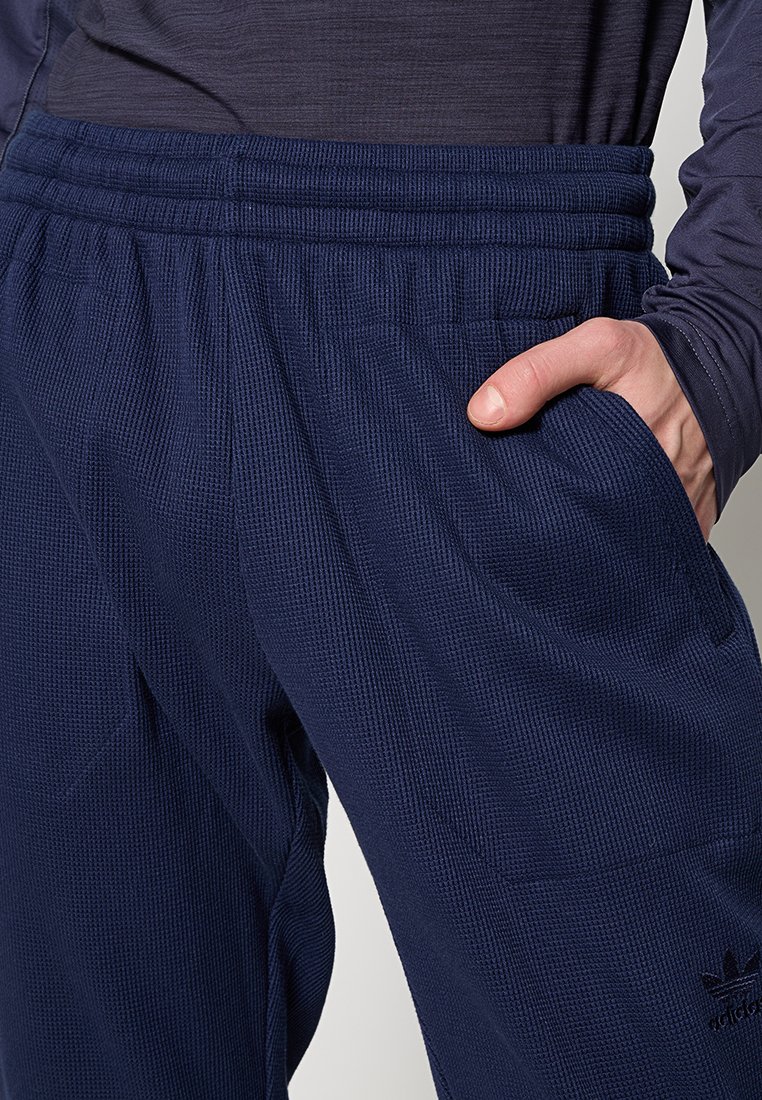 Marineblauwe sportbroek met een gestructureerde stof, elastische tailleband en zijzakken; heeft een logo aan de onderzijde.