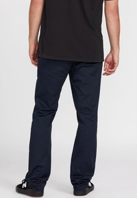 Navyblaue Hosen aus einem glatten Stoff, mit geradem Schnitt und minimaler Nahtverarbeitung. Getragen mit einem schwarzen T-Shirt und schwarzen Schuhen.