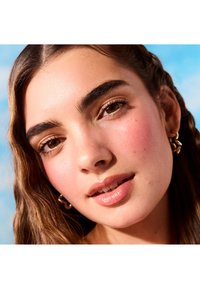 Nahaufnahme eines Gesichts mit hellbraunen, lockigen Haaren. Zu den Merkmalen gehören goldene Ohrringe, natürliche Make-up und eine klare Haut mit einem zarten Hauch von Rouge.