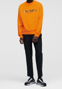 Oranssi crewneck-huppari, jossa on mustaa painettua tekstiä, yhdistettynä mustiin kapeneviin housuihin ja musta-valkoisiin lenkkareihin. Sileä pinta, rento istuvuus.