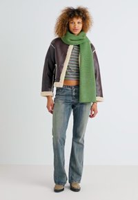 Veste en cuir marron avec doublure en fourrure synthétique, top court rayé, écharpe verte à texture, jean bleu évasé et chaussures beiges.