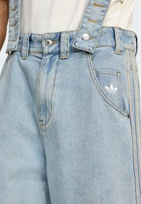 Svetlo modré džínsové overall s nastaviteľnými trakovými ramienkami, s gombíkovým uzáverom, detailným prešívaním a malým bielym logom na prednej vrecku.