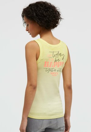 Frau mit lockigem Haar trägt ein ärmelloses gelbes Tanktop mit dem Text "Today be happy" auf der Rückseite, kombiniert mit grauen Jeans.