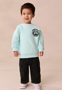 Jasnoniebieska bluza z napisem "Potwory i spółka" na rękawie, zestawiona z czarnymi spodniami cargo i białymi sneakersami. Gładki materiał, luźny krój.