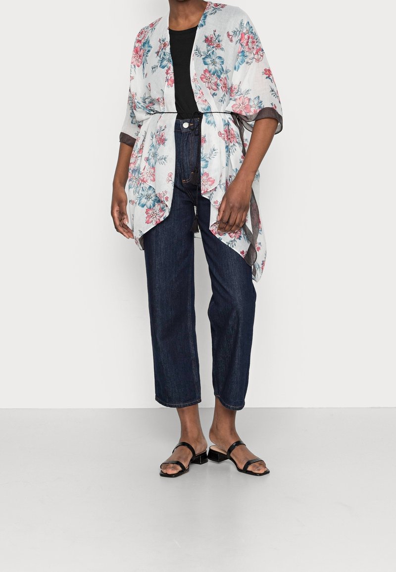 Personne portant un kimono blanc fleuri par-dessus une chemise noire, un jean bleu foncé et des sandales noires à talons bas, debout devant un fond clair uni.