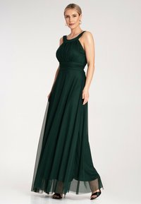 Sukienka maxi w głębokiej zieleni z plisowanym gorsetem, szerokimi ramiączkami i zwiewną spódnicą z przezroczystą nakładką. Akcentowana czarnymi szpilkami.