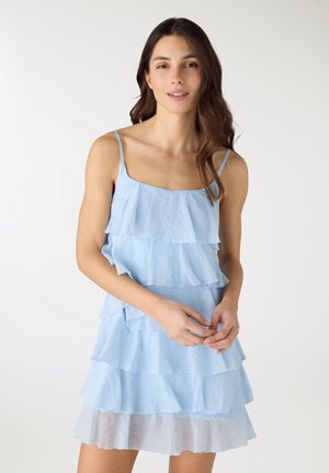 Giovane donna con lunghi capelli castani che indossa un vestito azzurro a balze senza maniche, in piedi davanti a uno sfondo bianco.