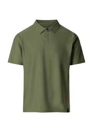 Uomo in piedi che indossa una polo verde oliva a maniche corte, pantaloni beige chiaro, scarpe da ginnastica bianche e tatuaggi visibili sulle braccia.