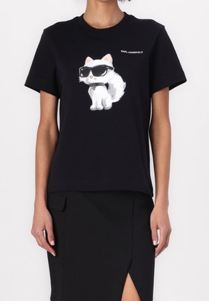 Osoba nosząca czarną koszulkę Karl Lagerfeld z białym nadrukiem kota w czarnych okularach przeciwsłonecznych oraz czarną spódnicę z przednim rozcięciem.
