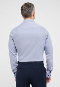 Camicia a righe leggera blu e bianca con maniche lunghe, polsini con bottoni e colletto standard, abbinata a pantaloni blu navy, vista da dietro.