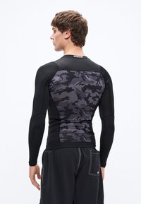 Czarna sportowa koszulka z długim rękawem z ciemnym wzorem kamuflażu na plecach. Posiada dopasowany krój i płaskie szwy dla komfortu.