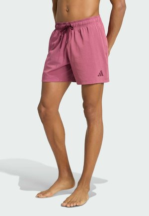 Persona scalza che indossa pantaloncini sportivi rosa con coulisse e piccolo logo sulla coscia sinistra, in piedi contro uno sfondo semplice.